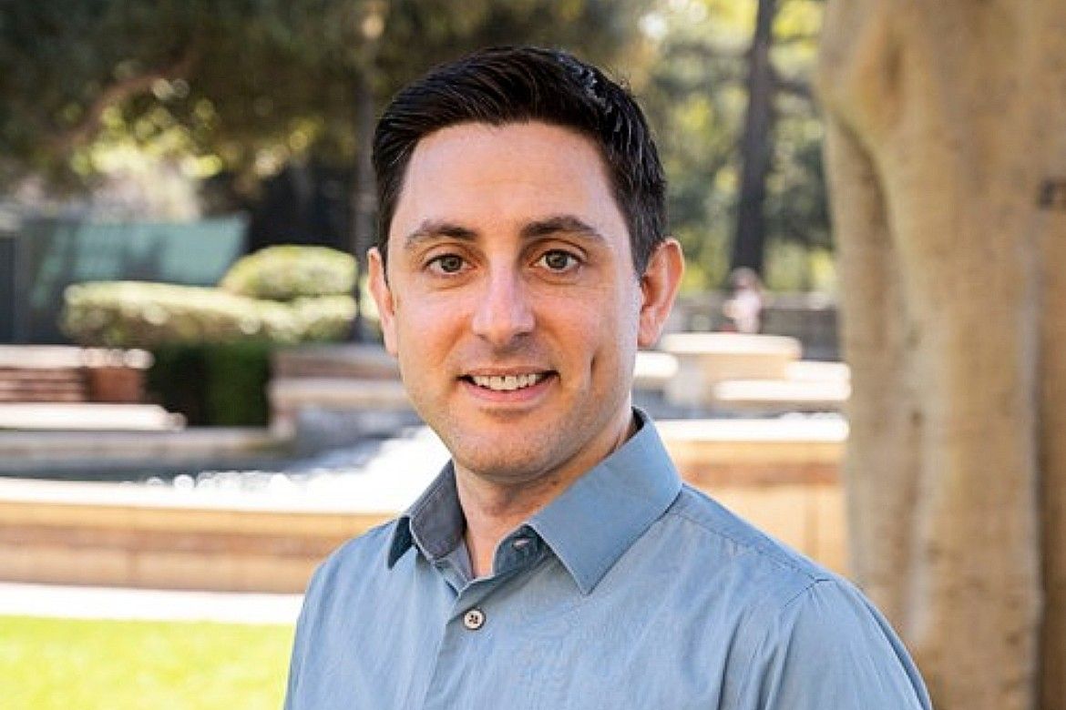 Headshot of Derek Schaffer, a man with short dark hair wearing a light blue shirt, smiling outdoors on a sunny campus setting with trees and a fountain in the background.