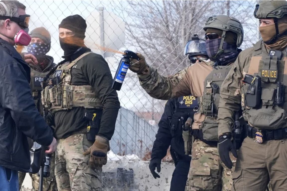 Armed law enforcement officers in tactical gear and face coverings, with one officer deploying pepper spray, standing near a chain-link fence in a winter setting