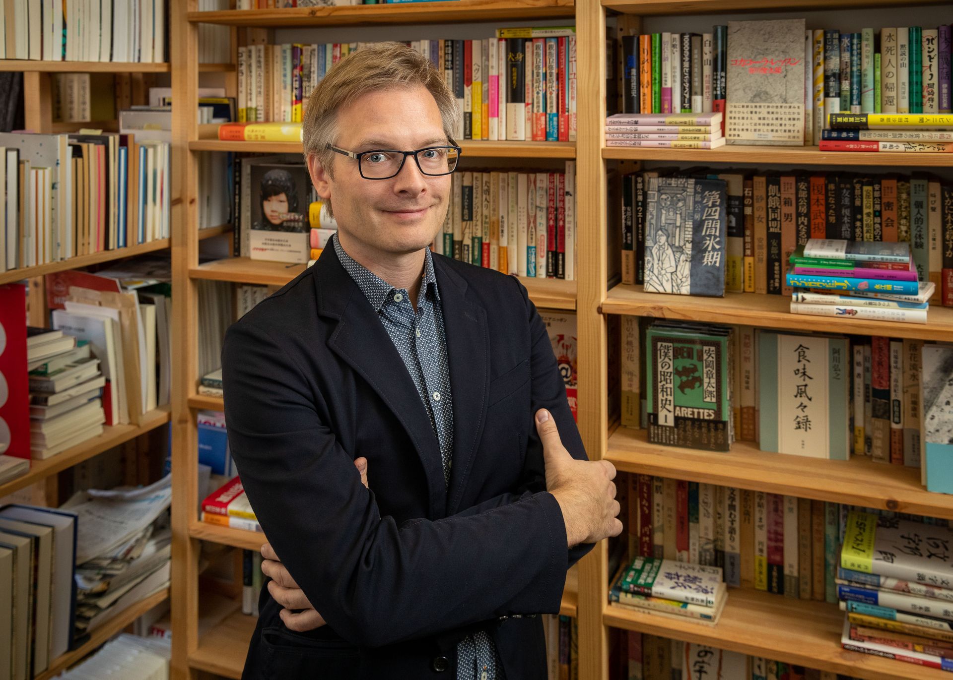 Michael Emmerich, in black jacket and glasses, stands with arms crossed in front of bookshelves full of Japanese books