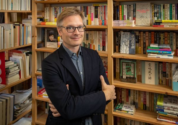 Michael Emmerich, in black jacket and glasses, stands with arms crossed in front of bookshelves full of Japanese books