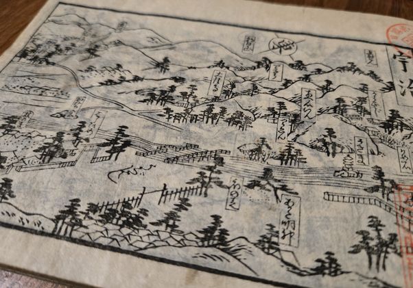 A historical Japanese woodblock-printed map showing mountains, trees, roads, and labeled landmarks with Japanese text, printed on aged paper with red seal stamps visible on the right edge.