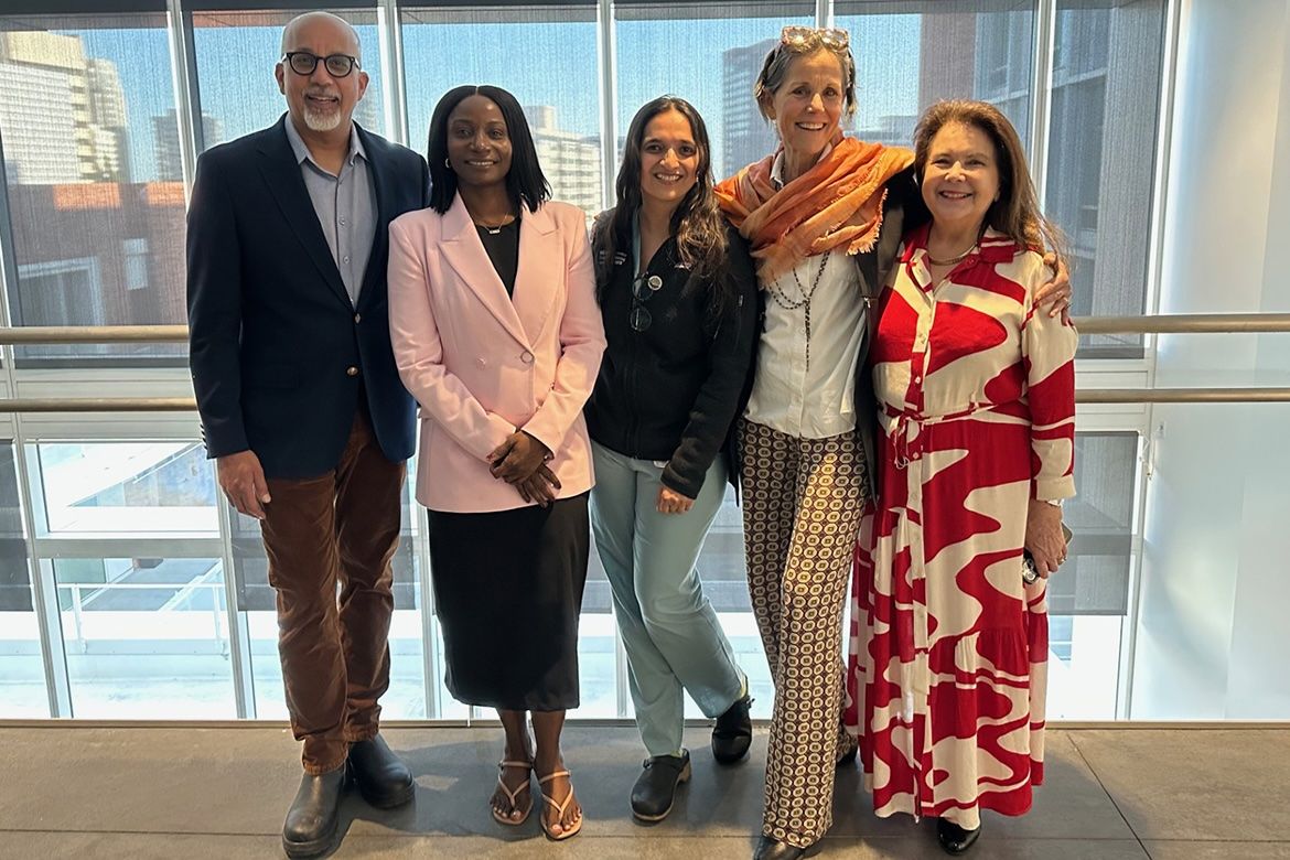 From left to right, Dr. Sadath Sayeed, Fatou Baldeh, Dr. Aparna Sridhar, Dr. Wendelin Slusser, and Mary Machado Schammel stand smiling together for a group portrait in front of a large window at UCLA.