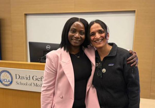 Fatou Baldeh, in a pink blazer, and Dr. Aparna Sridhar, in a black zip-up jacket, stand with their arms around each other smiling in front of a David Geffen School of Medicine at UCLA podium.