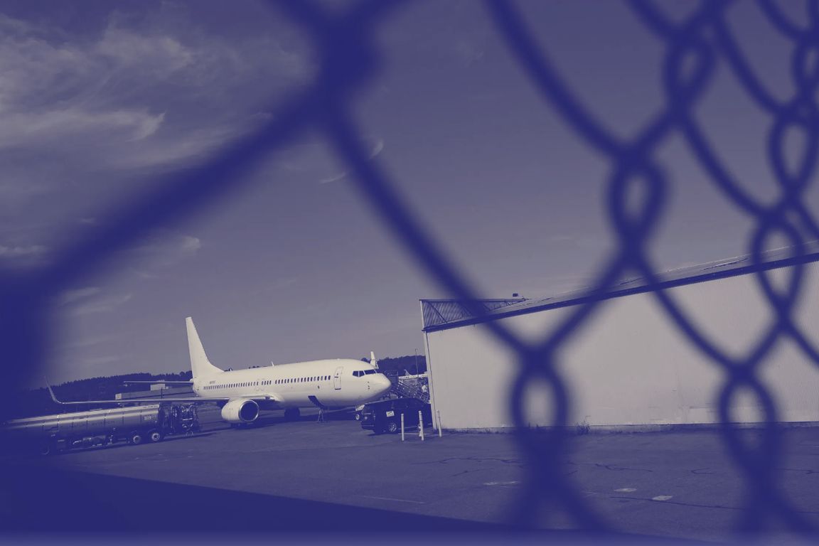 A commercial airplane parked on a tarmac next to a hangar, photographed through a chain-link fence with a blue-tinted filter.