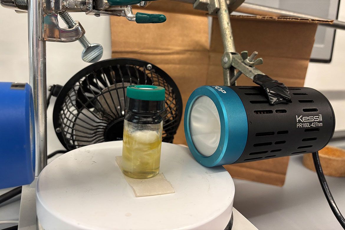 A photochemical reaction setup with a small vial containing a yellow solution on a white platform, illuminated by a Kessil PR160L 427nm light, with a small fan in the background for cooling.