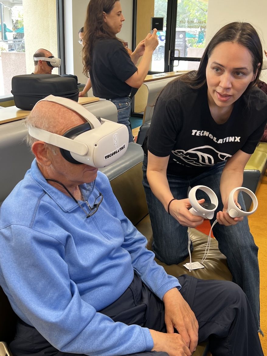 An older man wearing a virtual reality headset sits while a young woman from the UCLA TecnoLatinx group guides him through a VR tutorial, holding the controllers beside him.