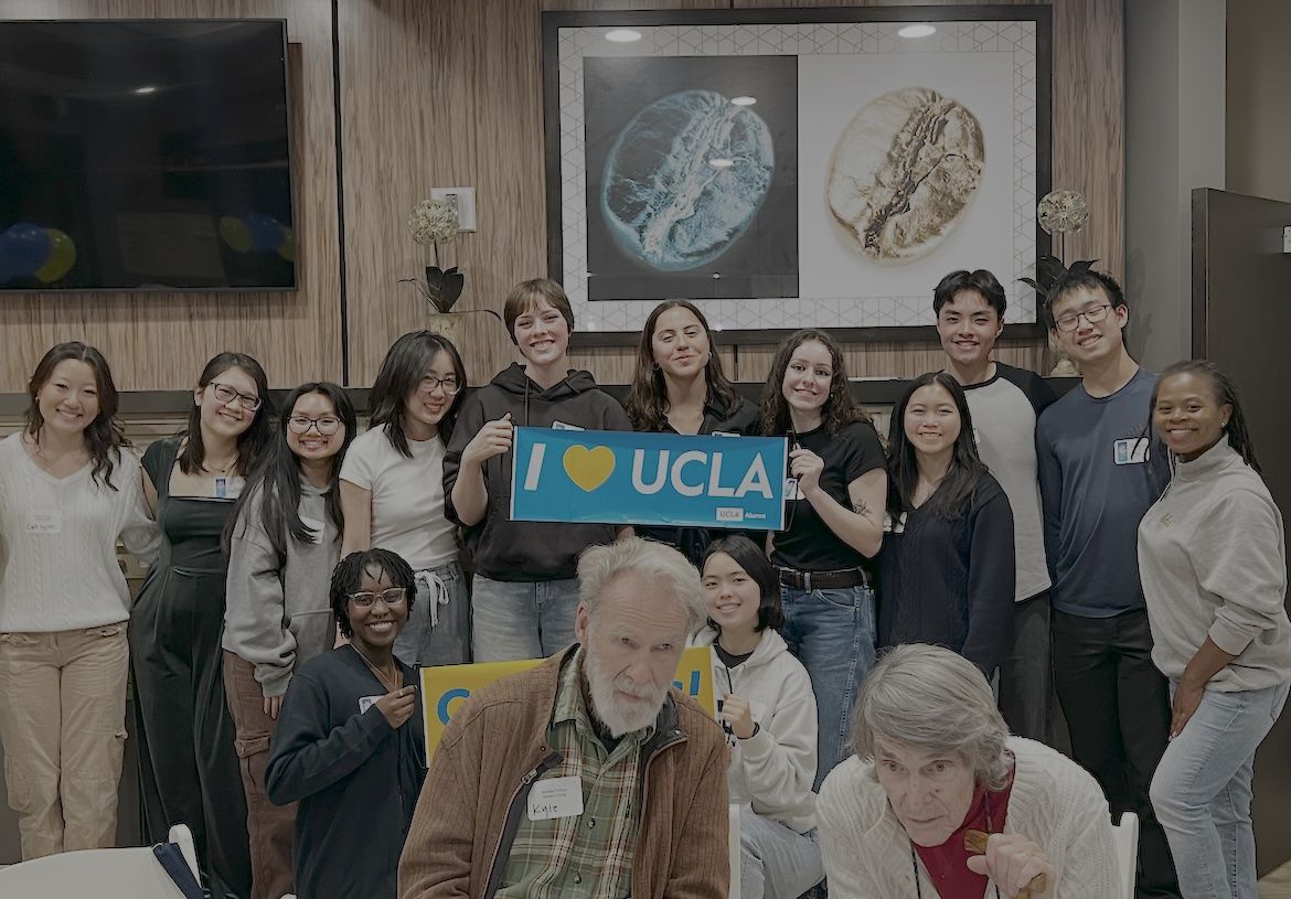 Students and older adults gather at a dining table at Belmont Village Senior Living during a Dinner for 12 Strangers event, smiling and holding a blue “I love UCLA” sign, as they build community and share stories across generations.