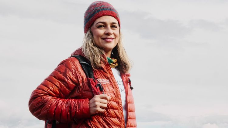 Sara Safari smiles outdoors wearing a red puffer jacket and striped knit hat against an overcast sky.
