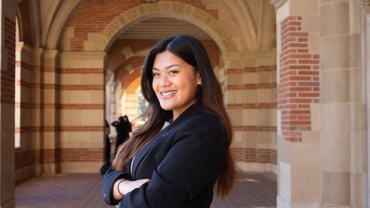 UCLA student Sheryl Samala stands with arms crossed and smiles in a black blazer beneath the red brick arches of a UCLA campus building.
