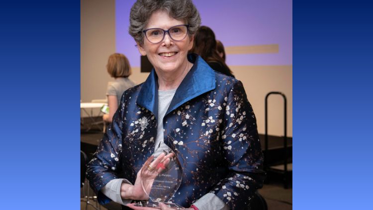 Barbra Nelson smiles while holding a glass Torchbearer Award trophy, wearing a floral brocade jacket at an awards ceremony.