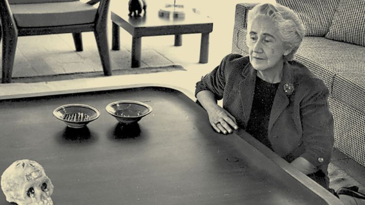 A black-and-white archival photo of designer Clara Porset seated at a low table with two decorative bowls and a skull, with mid-century modern furniture visible in the background.