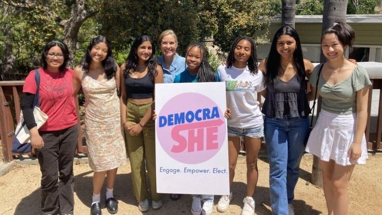 A group of eight young women stand together outdoors holding a pink and blue "DemocraShe: Engage. Empower. Elect." sign.