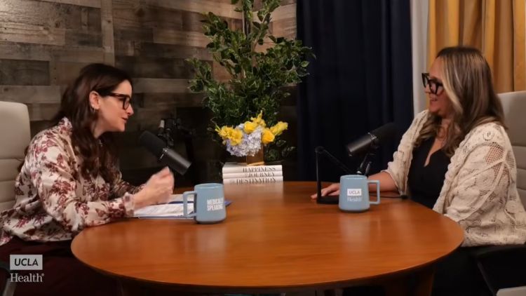 Two women wearing glasses sit across from each other at a round table with microphones and UCLA Health mugs during a taping of the "Medically Speaking" podcast.