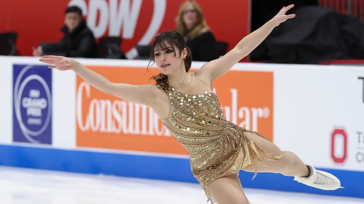 Figure skater Alysa Liu performs on the ice in a gold sequined dress with arms outstretched.