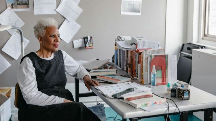 Nell Irvin Painter sits at a desk covered with books and papers, gazing thoughtfully to the side in her office.