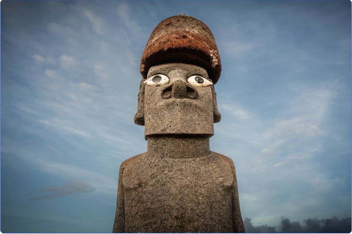 A close-up of a moai stone statue on Easter Island with white coral eyes and a red stone hat, photographed against a blue cloudy sky.