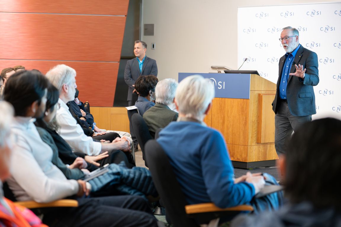 Fred Ramsdell speaks at a CNSI podium before a seated audience in an auditorium, while Hector Aguilar-Carreno stands in the background near the wall.