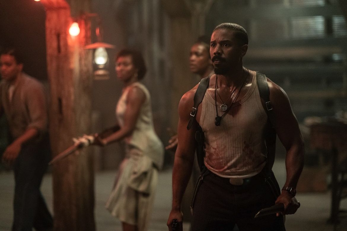Actor Michael B. Jordan, with blood on his shirt and a weapon in hand, stands alert in a tense, dimly lit scene from the film "Sinners," with other characters visible in the background. Photo courtesy Warner Bros.