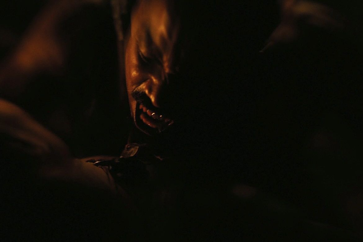A close-up of a vampire's face with bared fangs in a dark, atmospheric scene from the film "Sinners.