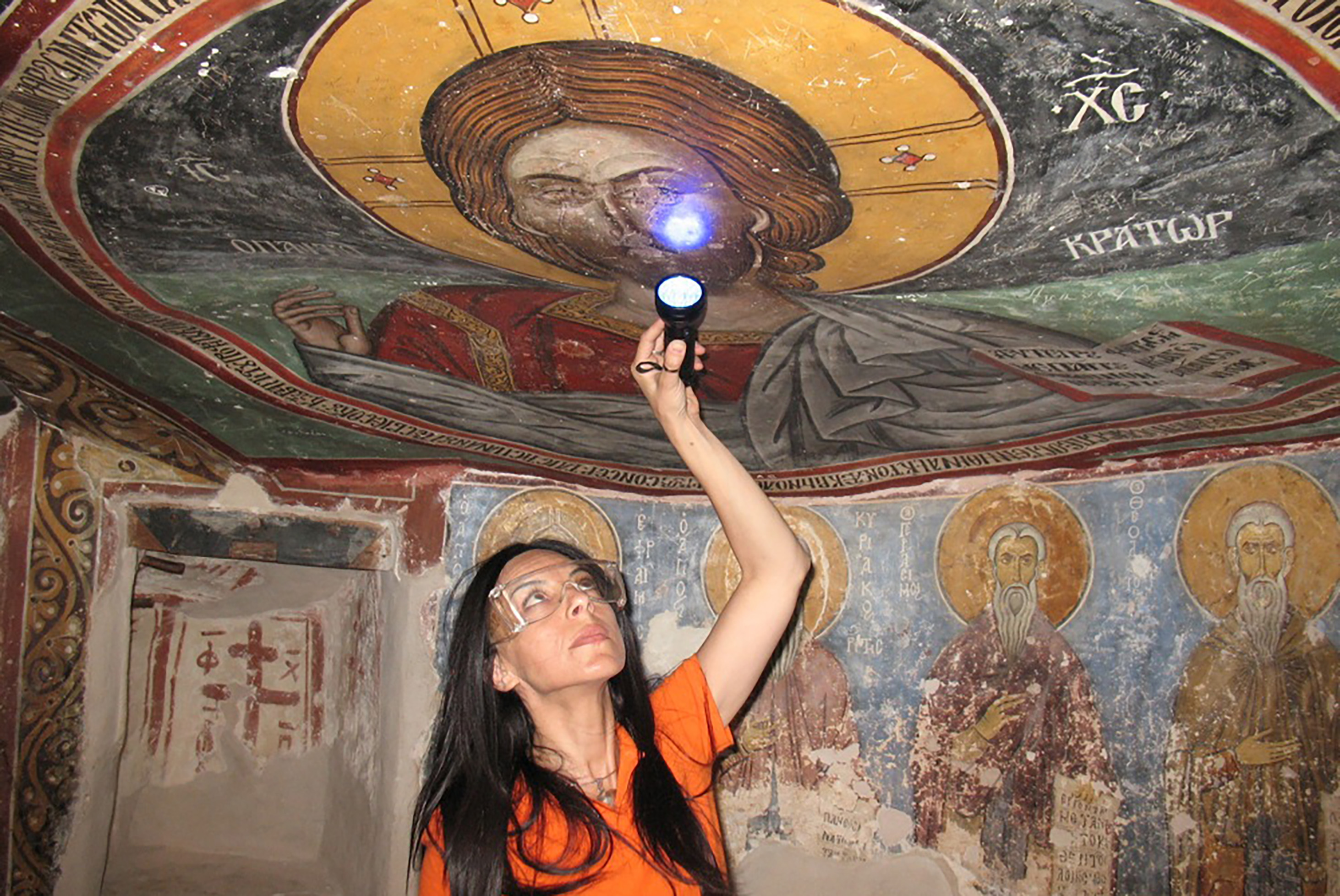Ioanna Kakoulli examining Byzantine wall paintings with a UV light in the cave church of St. Neophytos Monastery near Paphos, Cyprus, with religious frescoes visible on the surrounding walls and ceiling.