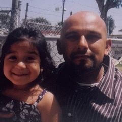 A childhood photo of a young Cianna Ulloa smiling alongside her father Cesar Ulloa, with a chain-link fence and trees visible in the background.