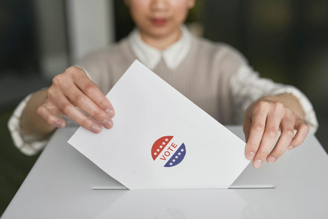 A person slides a ballot envelope marked with a red, white and blue "Vote" sticker into a ballot box.