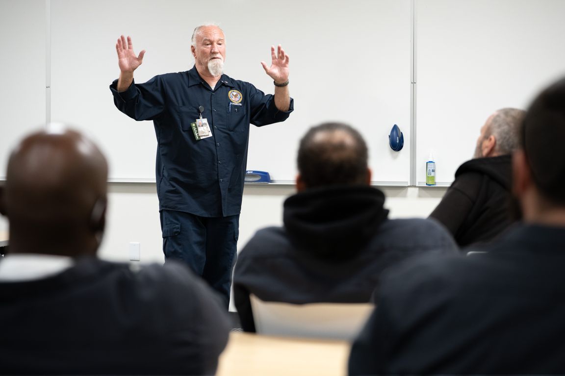 Daniel Martin, wearing a navy uniform and badge, stands at the front of a classroom with hands raised while addressing a group of seated students at the Veterans Success Academy at UCLA Extension.