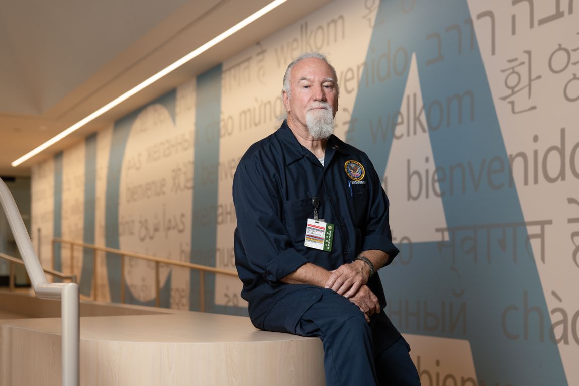 Daniel Martin sits in front of a large UCLA mural featuring welcome greetings in multiple languages, wearing his navy uniform and badge.