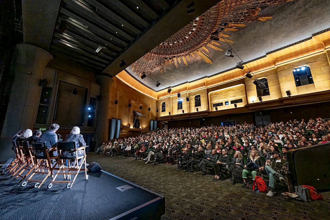 Two costume designers sit on director's chairs on a stage at the front of a packed ornate theater, addressing a large audience seated in the orchestra and balcony levels.