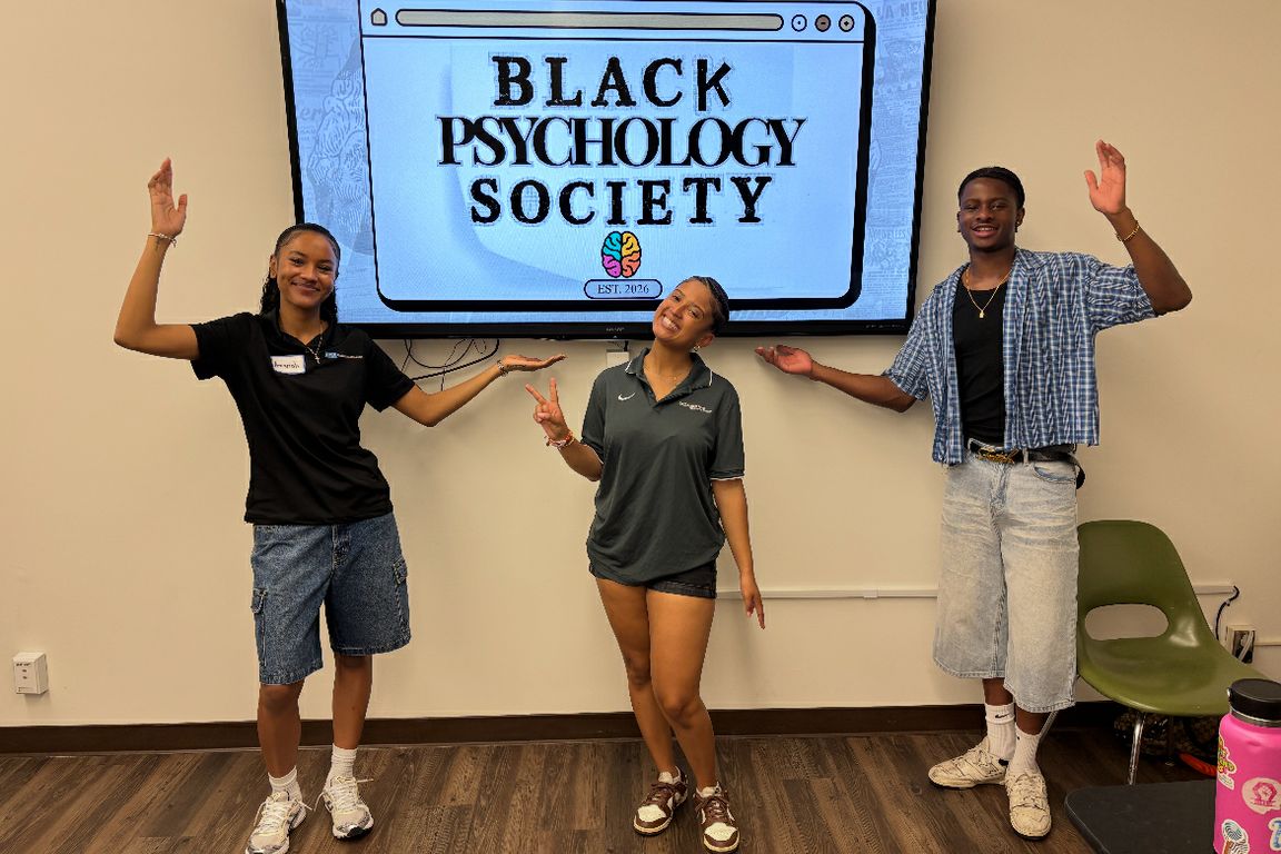 Three students strike playful poses in front of a large screen displaying the Black Psychology Society logo in a campus meeting room.