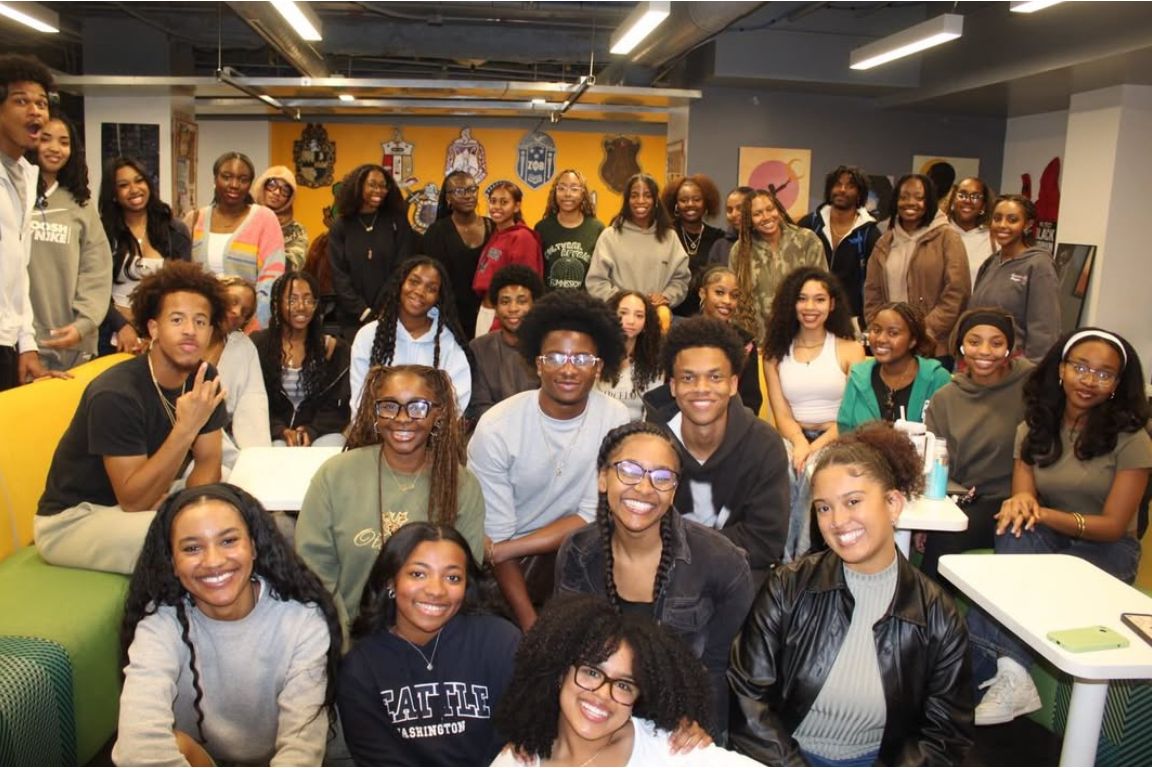A large group of smiling students crowd together for a photo inside the Black Bruin Resource Center, with the center's colorful yellow wall and fraternity and sorority crests visible in the background.