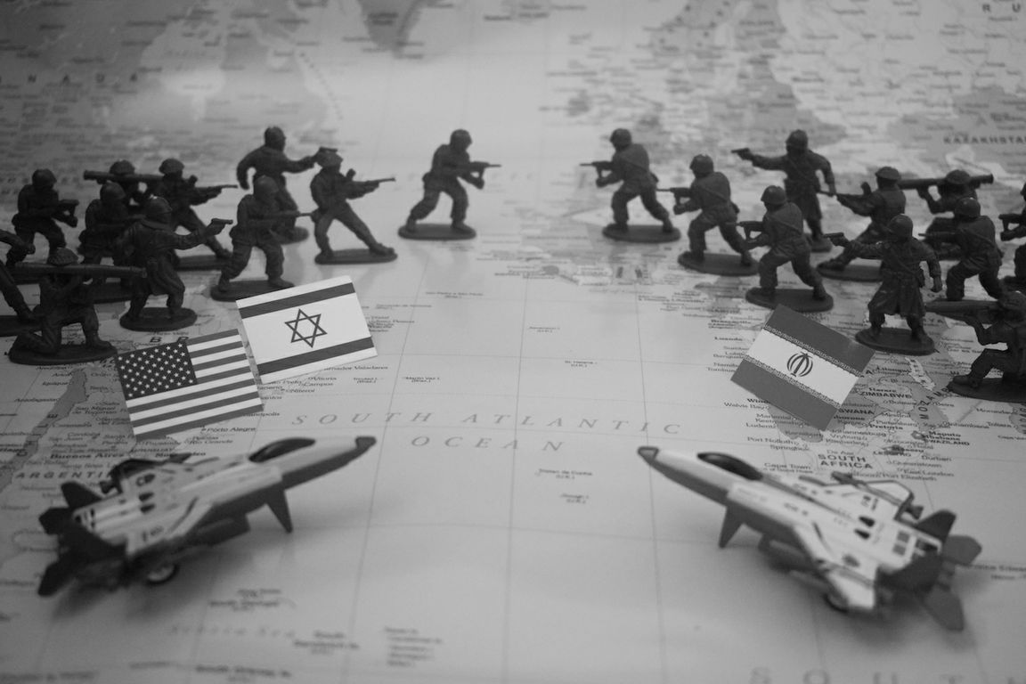 A black-and-white conceptual illustration shows two opposing groups of toy soldiers facing each other on a world map, with miniature flags of the United States and Israel on one side and Iran on the other, flanked by model fighter jets.