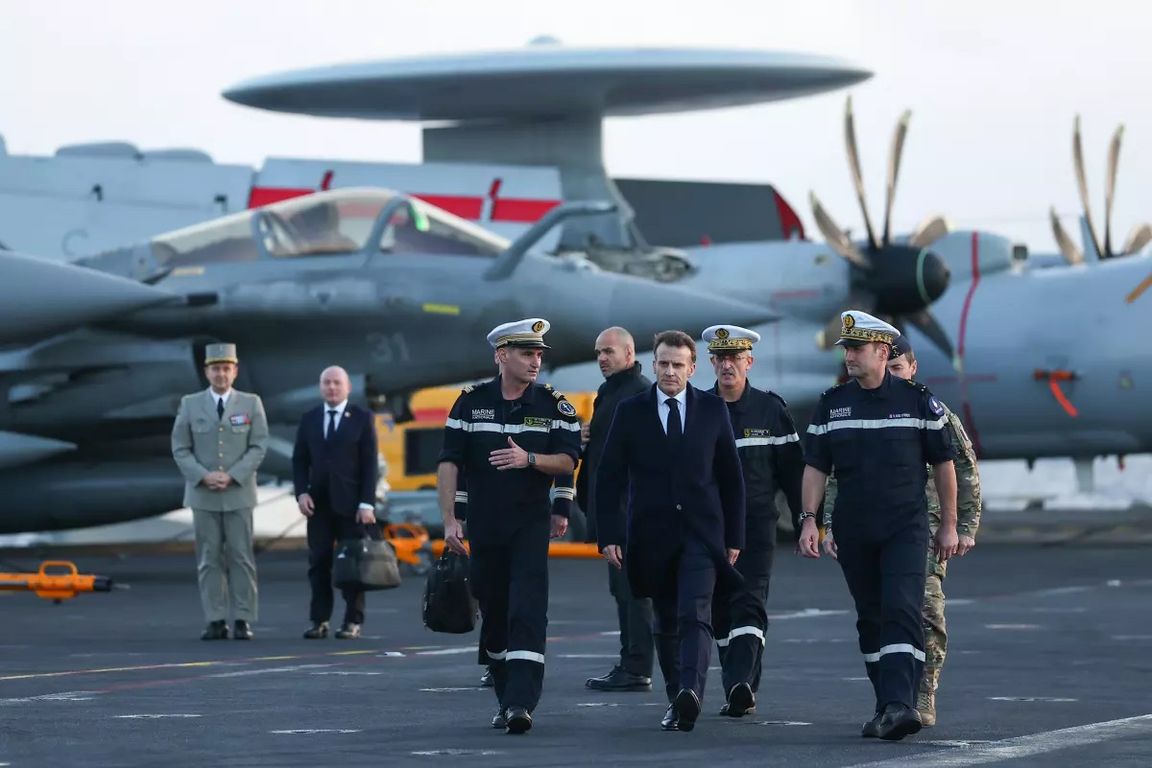 French President Emmanuel Macron walks alongside French military officers on an aircraft carrier flight deck, with military aircraft visible in the background.