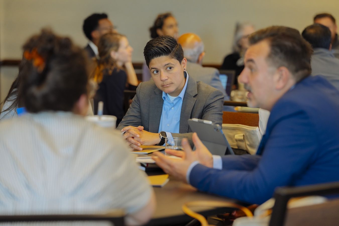 Conference attendees engaged in small group discussion at a table, with one person in a gray blazer listening attentively while another gestures while speaking.