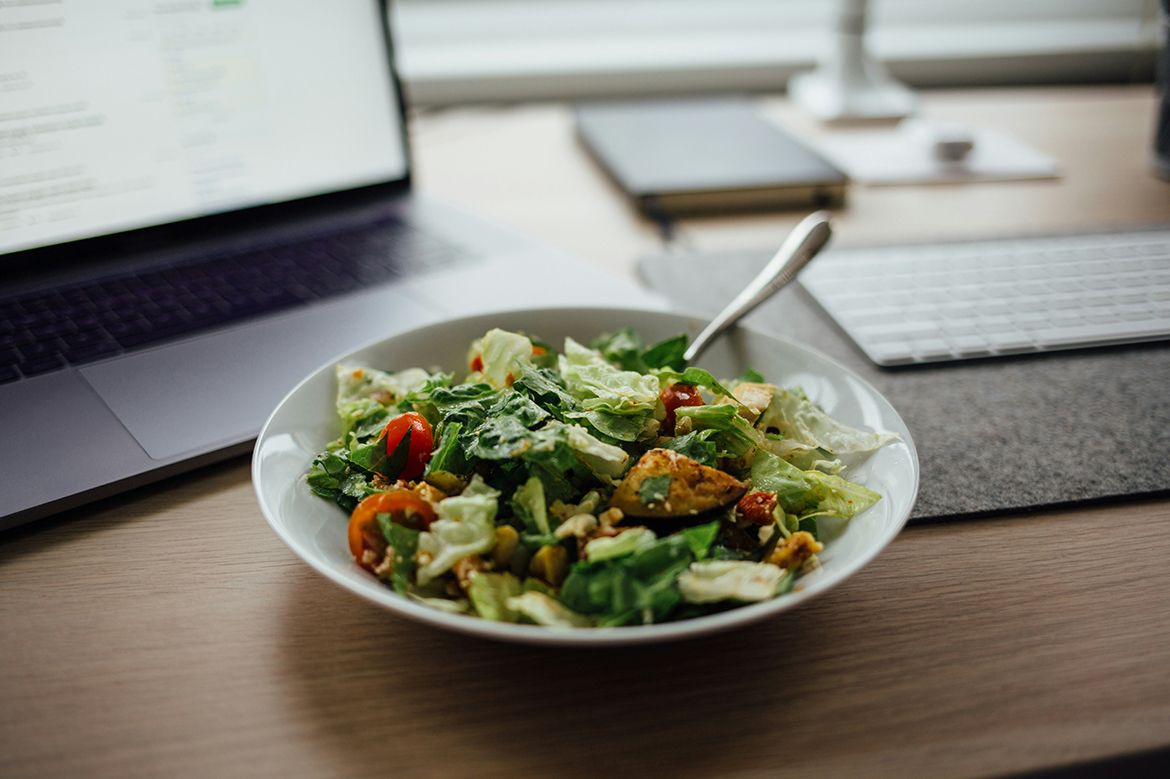 A fresh green salad with cherry tomatoes and croutons sits on a desk beside a laptop and keyboard.