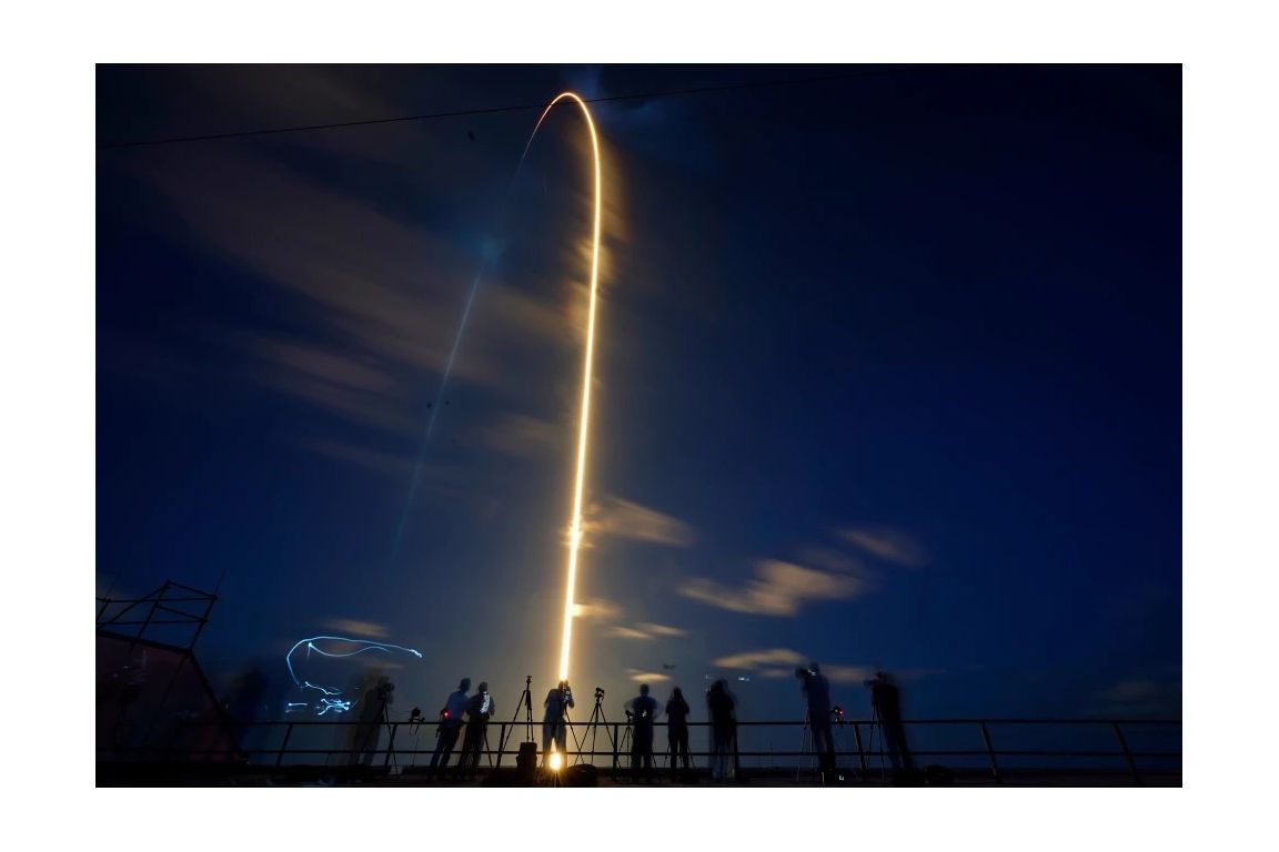 Silhouetted spectators and photographers watch as a rocket traces a bright arc across a deep blue night sky during a launch.