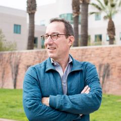 A headshot of David Eisenman smiling outdoors with arms crossed, with palm trees and a building visible in the background.