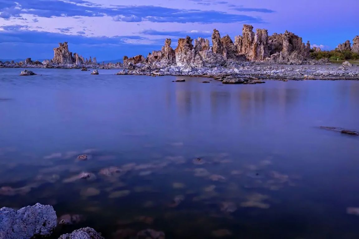 The distinctive tufa rock formations of Mono Lake rise from the calm, glassy water at dusk under a blue and purple sky.