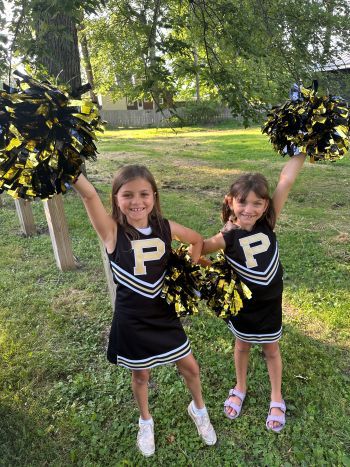 Two young girls smile and wave black and gold pom-poms while wearing matching Purdue cheerleading uniforms outdoors.