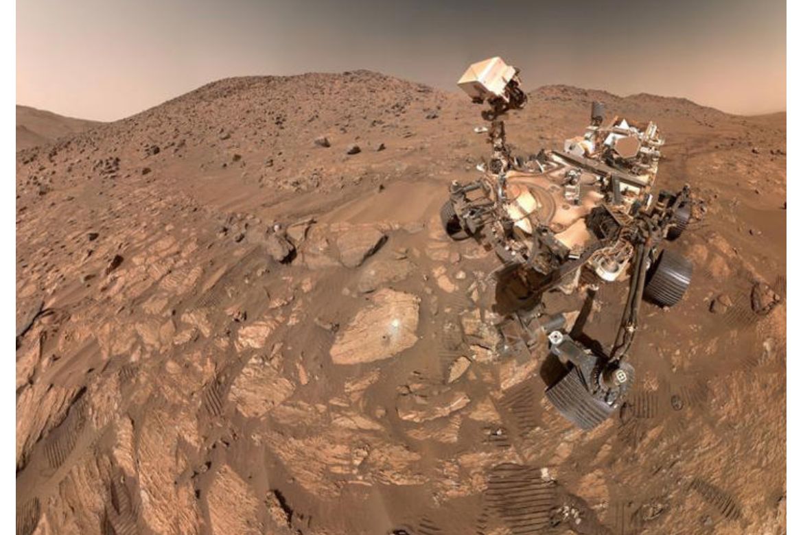 NASA's Perseverance rover captured in a self-portrait on the rocky, rust-colored surface of Mars, with rolling terrain visible in the background.