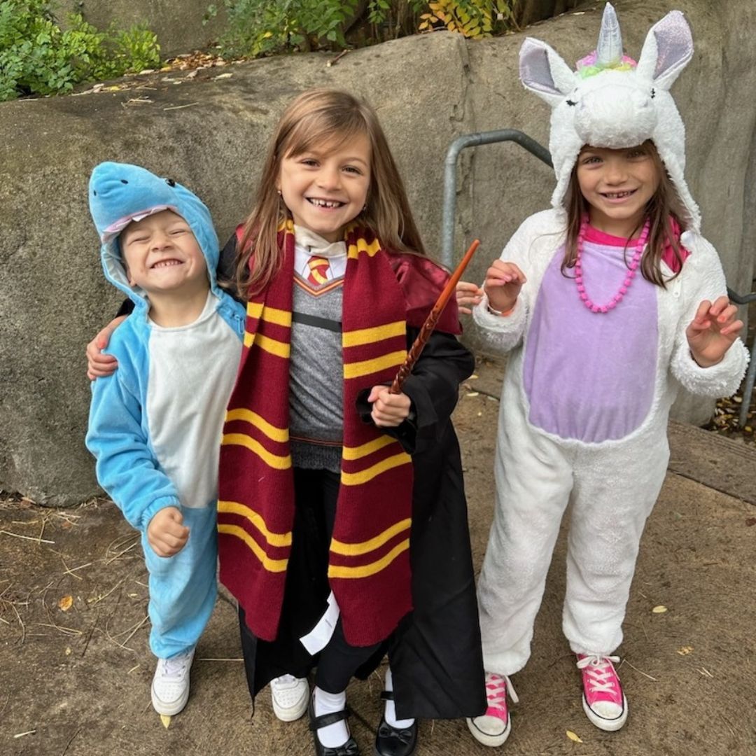 Three young children smile together in Halloween costumes, dressed as a shark, a Hogwarts student and a unicorn.