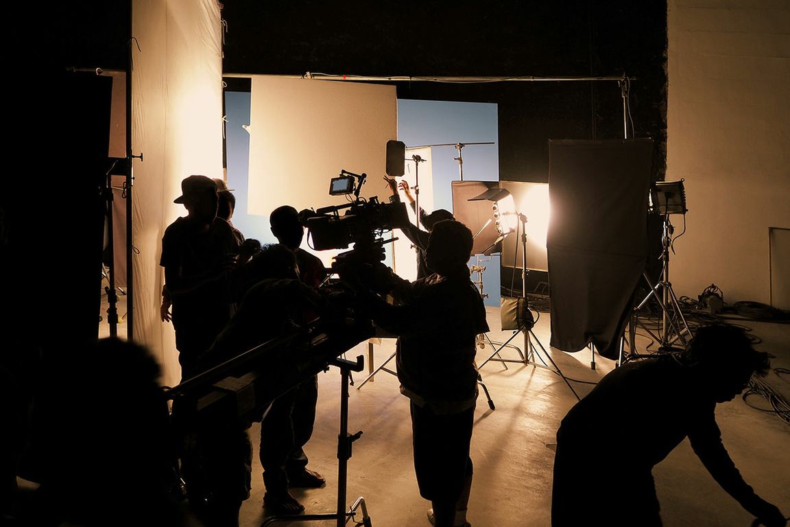 Silhouetted film crew members operate a camera and adjust lighting equipment on a production set.