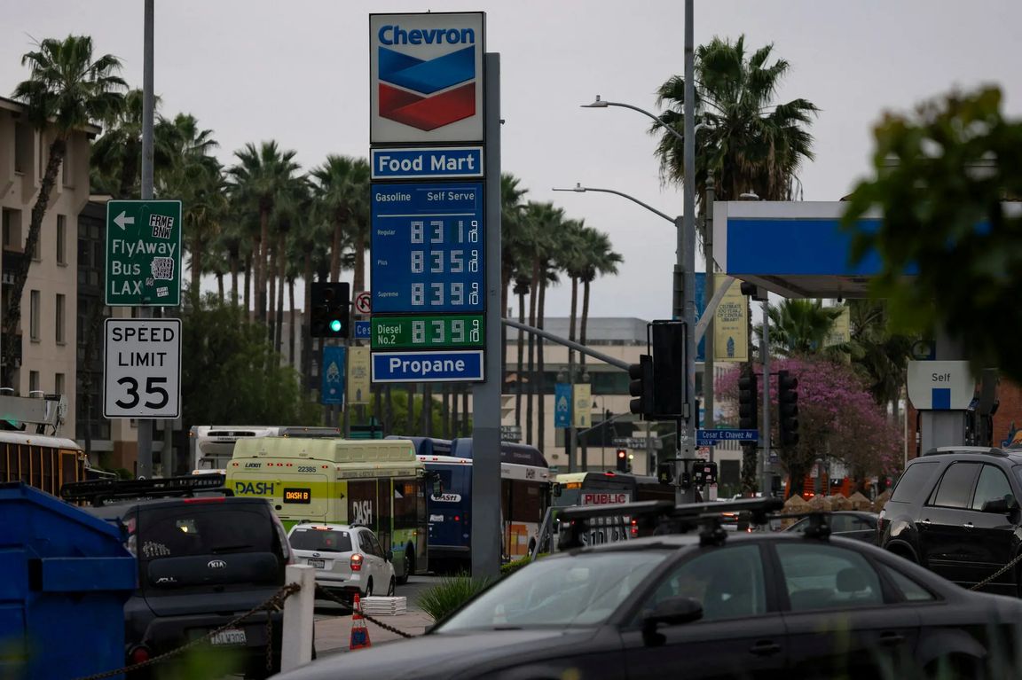 A Chevron gas station sign displays prices above $8 per gallon along a busy Los Angeles street lined with palm trees, traffic, and signs for LAX and a 35 mph speed limit.