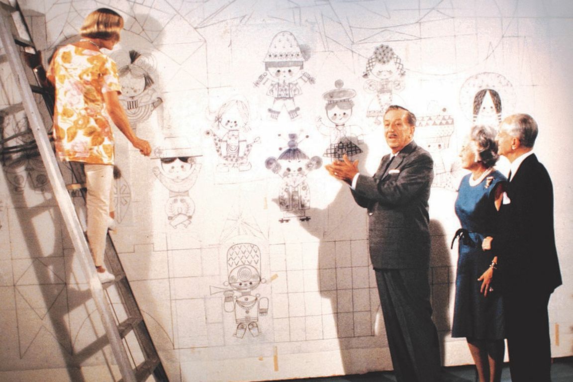 Artist Mary Blair works on a large illustrated mural from atop a ladder while Walt Disney and Doris and Jules Stein look on in an archival photograph.