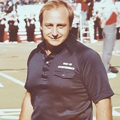Mazzone at UCLA A young Angelo Mazzone III stands on a football field sideline wearing a dark Pac-10 Conference polo shirt, with a marching band visible in the background.