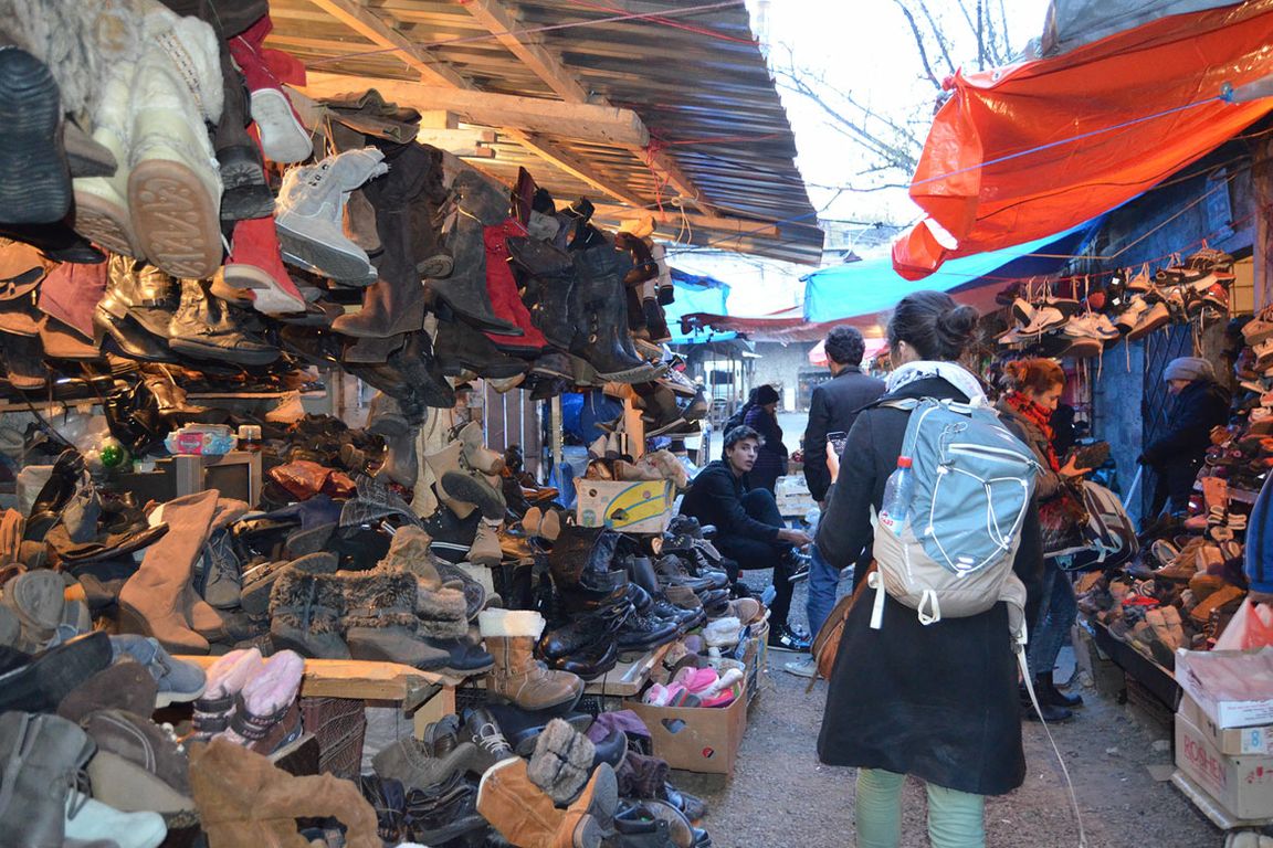 Shoppers browse a crowded outdoor market stall overflowing with used shoes and boots hanging overhead and stacked on the ground.