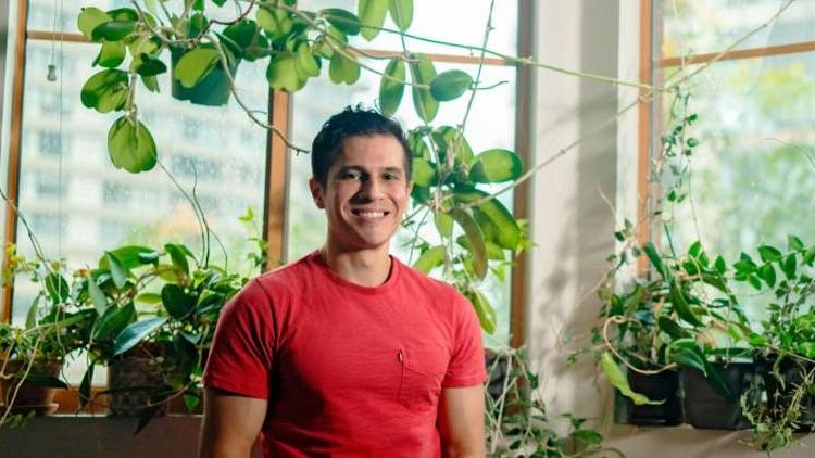 Dr. Nasser S. El-Okdi smiles while seated in front of a sunlit window surrounded by lush green houseplants.