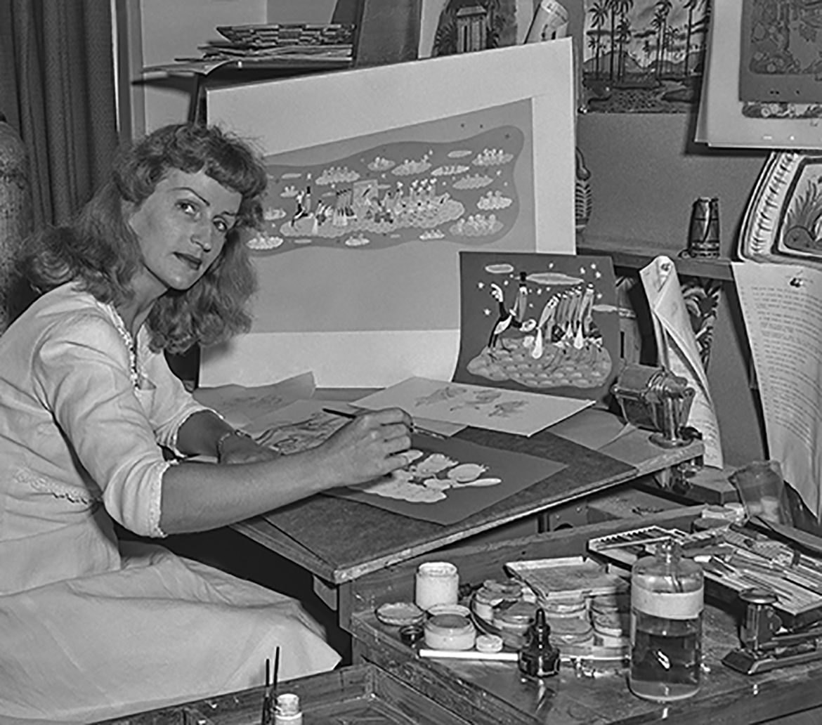 A black-and-white archival photograph from 1943 shows Disney Legend Mary Blair seated at her art desk, designing colorful illustrations for St. Joseph's Hospital in California, with paint supplies and artwork visible around her.