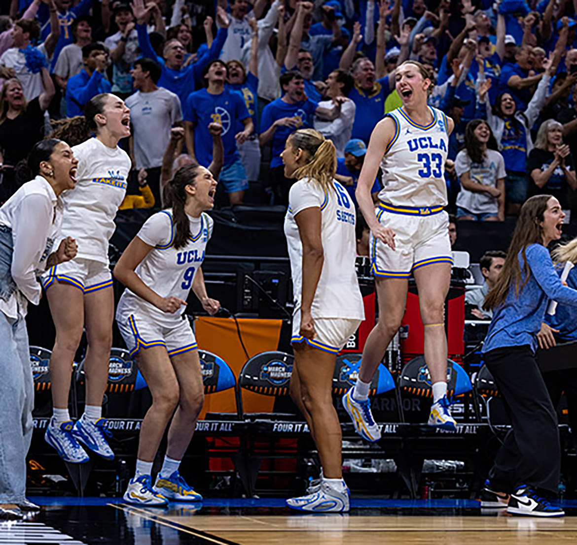 UCLA Bruins players and staff leap and cheer from the bench as the team clinches their Elite Eight victory, with fans celebrating in the packed arena behind them.