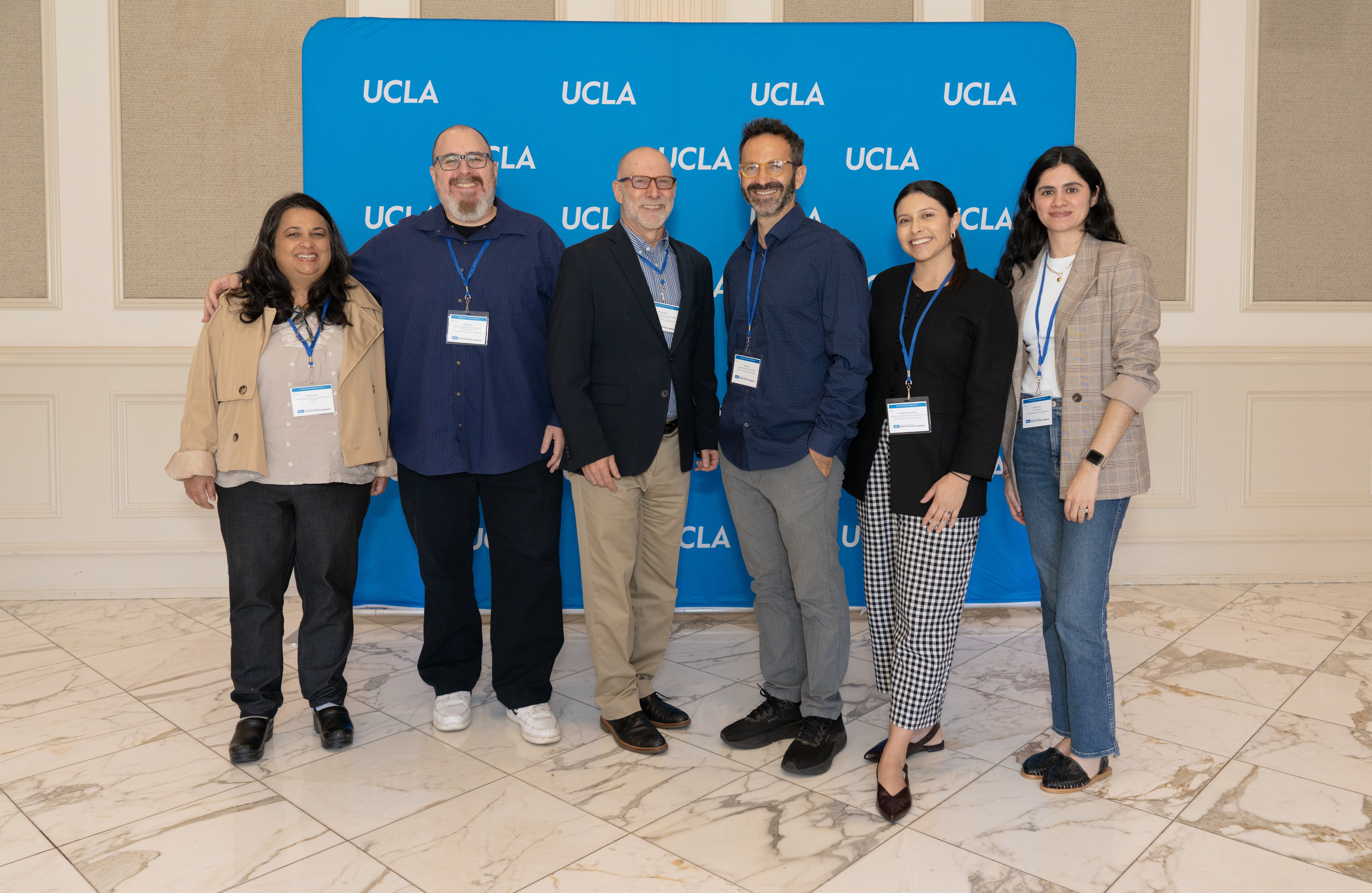 Six people smile and pose together in front of a blue UCLA step-and-repeat banner at a professional event. All six wear conference lanyards with name badges. The group includes three men and three women standing in a row on a marble floor inside a formal venue.